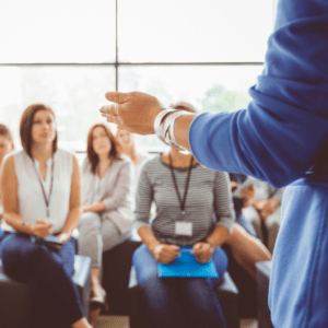 A speaker gestures while addressing a group of attentive professionals seated in a bright, modern training room.