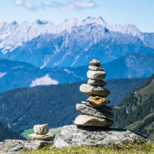 Stacked stones on a mountain ridge with a backdrop of lush green hills and majestic snow-capped peaks under a bright blue sky.