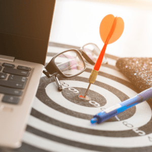 A dart hitting the bullseye on a target board, placed next to a laptop, glasses, and a pen, symbolizing goal setting, focus, and achieving success.